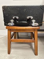 Front view of closed black metal carrying case on stool, showing handle and latches