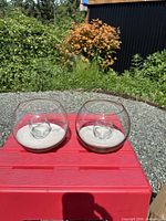 Two glass fishbowl candle holders on red table, outdoor setting