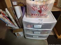 Plastic three-drawer organizer with bag of gift bags on top and box of assorted wrapping paper rolls beside it