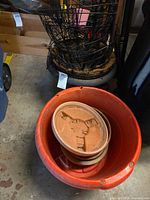 Stack of wire hanging baskets in black planter next to orange bucket containing terracotta saucers