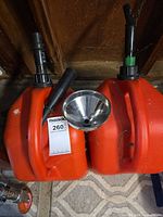 Two red-orange plastic 5-gallon gas cans with spouts, stainless funnel on top