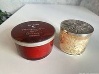 Two candles side by side, red tin and cream jar