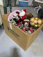 Cardboard box containing assorted Christmas decorations including plush Santa, gold ball ornaments, pinecones and other items