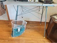 Full view of ironing board standing on hardwood floor, teal afghan in clear bag beneath