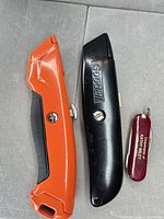 Three cutting tools laid out: orange utility knife, black Goodell knife, red pocket multi-tool