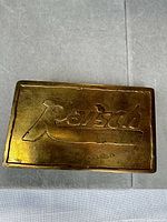 Front of rectangular brass-tone buckle showing engraved "Rausch" logo