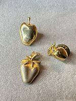 Group shot of all three goldtone fruit pieces (apple brooch, strawberry pendant, apple lapel pin) on grey background