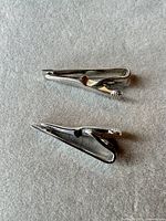 Two silver-tone tie clips on grey background, side view highlighting overall shape.