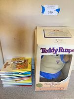 Front view showing Teddy Ruxpin plush inside box next to stack of books and cassettes