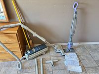 Full lot showing Electrolux vacuum, hose, wands, powerhead, crevice tool, Shark steam mop, replacement pads, straw broom, spare handle