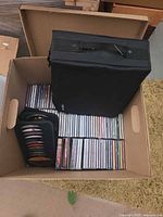 Box containing rows of jewel case CDs, large black CD binder on top, small CD wallet at side