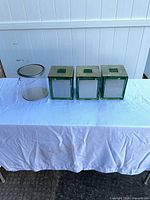 Full view of three green glass canisters with metal lids and one clear plastic pail with lid and handle on table