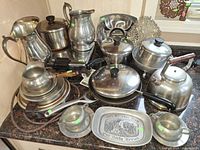 Group shot of all metal cookware, pitchers, kettle, warming bases and trivet