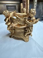 Angled view of gilded candle holder showing three cherubs around ornate base