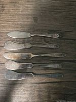 Five matching silver-plated butter spreaders laid out to show length, blade shape and handle pattern