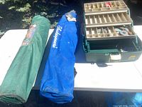 Two camp chairs in carry bags (green, blue Shakespeare) beside Plano tackle box on table