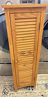 Front view of tall narrow wood cabinet with louvered door and knob