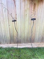 Two metal garden stakes leaning against fence showing full height and candle holder tops with twisted wire