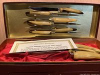 Interior of case showing five knives in lid, one knife on satin lining, guarantee certificate visible