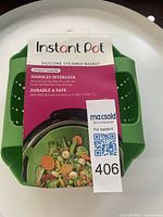 Retail-packaged green Instant Pot silicone steamer basket showing product information tag and QR label 406