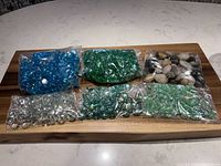 Six bags of assorted decorative glass gems and river rocks on wooden board