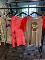 Three tops on hangers: gray Wicked hoodie, pink long-sleeve Under Armour tee, grey/red Wonder Woman Under Armour tee