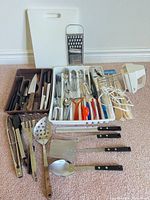 Overview of all kitchen items including cutting board, box grater, utensil trays, knives, flatware, serving tools, hand mixer