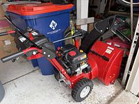 Overall view of red two-stage gas snow blower with black chute and control handles in garage