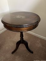 Round mahogany brown tripod pedestal side table showing top surface with stain and edge wear on carpeted floor