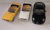 Front view of three 1:18 scale diecast cars: yellow Ford Mustang convertible, white Ford Mustang convertible, black Porsche 911 coupé