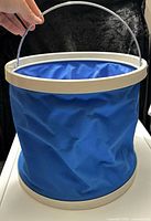 Side view of blue collapsible bucket showing nylon body, plastic rim/base, metal handle