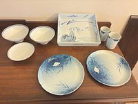 Group view of nine blue and white ceramic and porcelain pieces: two plates, three bowls, two cups, one square tray