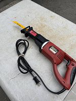 Full view of red corded reciprocating saw, attached yellow blade, bundled power cord