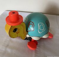 Left side view showing wooden head, red hat, turquoise shell, and red feet