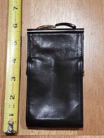 Front view of cigarette case next to ruler showing ~6 in height, black leather exterior, gold-tone clasp visible