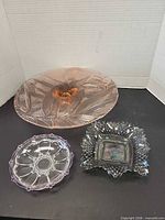 Group view of three glass pieces: pink cake plate, pink-tinted trinket dish, gray iridescent bowl