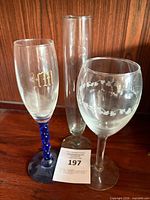 Three glass items grouped: wine glass with etched vine pattern, champagne flute with blue twisted stem and gold monogram, tall cylindrical vase