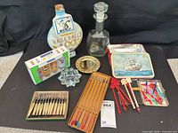Group shot of entire lot showing decanter, commemorative bottle, boxed Luminarc shot glasses, fondue forks, mini forks, cocktail picks, decorative tin, brass dish and glass star