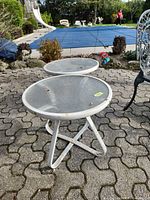 Pair of round white metal side tables with clear glass tops positioned outdoors