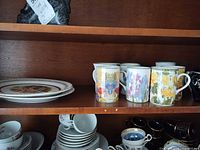 Stack of scenic dinner plates and group of floral mugs on shelf with additional teacups below