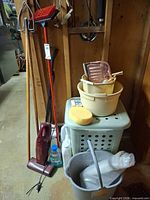 Overall view showing stick vacuum, hamper, buckets, mop handle, sponge, brush and cleaner bottle against wall