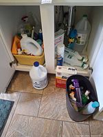 Overall view of open under-sink cabinet showing majority of cleaners, bleach jugs, bins