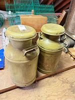 Two green painted milk cans side by side on wooden surface showing overall form, lids and handles