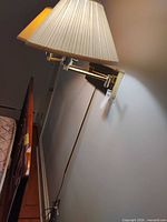 Side view of installed brass-tone swing arm wall lamp with pleated cream shade, visible cord and plug
