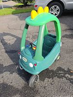 Front angled view of green ride-on car showing roof, dinosaur plates, wheels, steering wheel