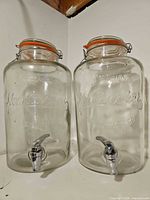 Front view of both glass dispensers showing embossed branding, lids and spigots