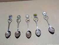 All five silver-plated souvenir spoons laid out bowl-down showing crests and charms
