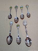 All seven silver plated souvenir teaspoons arranged on surface showing crests and bowls