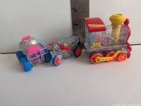 Front view of three clear plastic friction toys showing monster truck, motorcycle, train engine with ruler for scale