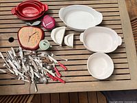 Overall lot showing flatware pile, red and white baking dishes, wall clock, mug, wedge dishes